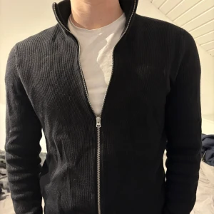 Svart ribbad tröja från Only & Sons - Säljer en stilren svart ribbad tröja från Only & Sons. Tröjan har en dragkedja framtill och är långärmad, perfekt för en avslappnad look. Passar bra till både jeans och chinos.