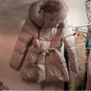  Beige dunjacka med pälsdetaljer från Fur Stockholm - Säljer en elegant beige dunjacka från Fur Stockholm. Jackan har en lyxig pälskantad huva och ett bälte i midjan för en snygg siluett. Perfekt för kalla dagar med sin varma och stilrena design.