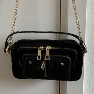Helena Recycled Nylon W. Gold - Snygg svart axelväska i skinn med guldfärgade kedjor och dragkedjor. Väskan har två framfickor med knappar och nitar på ovansidan för extra stil. Perfekt för att bära dina nödvändigheter med stil. Det var en julklapp som tyvärr inte passade.Helt ny.Oanvända. Ny pris 1029
