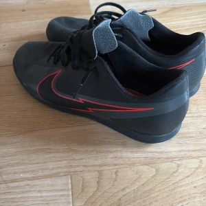 Svarta Nike fotbollsskor med röd detalj - Snygga svarta Nike fotbollsskor med en röd blixtdetalj på sidan. Skorna har snörning och en låg profil som ger en smidig passform. Perfekta för inomhusfotboll.