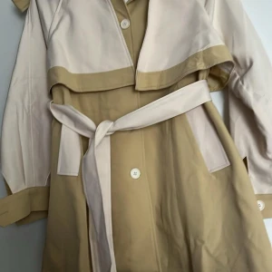 Beige och brun trenchcoat från Flo M Mode - Snygg trenchcoat i beige och brun från Flo M Mode. Jackan har långa ärmar och knyts med ett bälte i midjan. Den har knappar framtill och en klassisk krage. Perfekt för en stilren look.