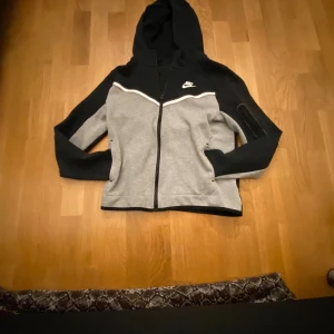 Grå och svart Nike tröja - Säljer en snygg grå och svart hoodie från Nike med dragkedja och huva. Den har en cool ficka med dragkedja på ärmen och Nike-loggan på bröstet. Perfekt för en casual look eller träning. 🖤