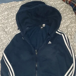 Mörkblå huvtröja från Adidas - Har fler bilder! Säljer en mörkblå huvtröja från Adidas med dragkedja och de klassiska tre ränderna i vitt längs ärmarna. Tröjan har en justerbar huva och är perfekt för en sportig look. Adidas-loggan är tryckt på bröstet. Perfekt för alla tillfällen! Pris kan diskuteras 