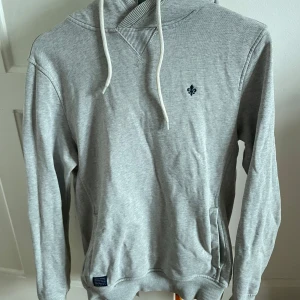 Grå hoodie från Morris - Säljer en stilren grå hoodie från Morris med en liten broderad detalj på bröstet. Tröjan har en klassisk passform med långa ärmar och en bekväm huva med dragsko. Perfekt för en avslappnad look.