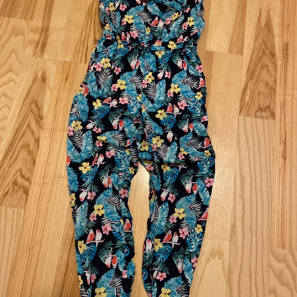 Säljer en färgglad byxdress med blommigt och papegojmönster. Den har smala axelband och en elastisk midja för en bekväm passform. Säljes i fint skick! . Farkut & Housut.