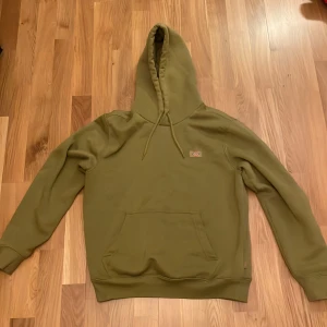 Olivgrön hoodie från Dickies - Säljer en snygg olivgrön hoodie från Dickies. Den har en klassisk design med en stor ficka fram och justerbar huva med snören. Perfekt för en avslappnad stil. Storlek XS herr, passar utan problem S (170cm~175cm).