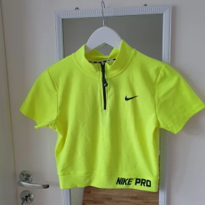 Neongul kortärmad topp från Nike - Säljer en neongul kortärmad topp från Nike med dragkedja framtill och Dri-FIT teknologi. Perfekt för träning med en sportig look och bekväm passform. Nike-logga på bröstet och texten 'NIKE PRO' vid nederkanten. 🏋️‍♀️