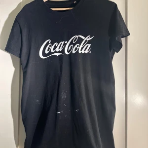 Svart Coca-Cola t-shirt - Säljer en svart t-shirt med Coca-Cola-logga i vitt. T-shirten har en klassisk passform och korta ärmar. Perfekt för dig som gillar retrostil och vill ha något bekvämt och stilrent i garderoben.