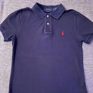 Mörkblå pikétröja från Ralph Lauren - Säljer en stilren mörkblå pikétröja från Ralph Lauren med klassisk krage och knappslå. Tröjan har en röd broderad logotyp på bröstet och korta ärmar. Perfekt för en familje event eller nyår  men ändå snygg look. Köpt för 800kr pris kan diskuteras