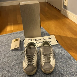 Golden Goose sneakers med gröna detaljer - Säljer ett par Golden Goose skor i mycket bra skick. De är vita med grå och gröna detaljer, och har en cool, sliten look. Passar till allt. Säljer dem för 3999. Originalförpackning och dustbag medföljer.