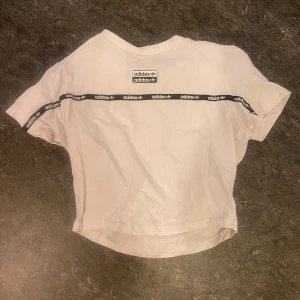 Vit t-shirt från Adidas - Säljer en snygg vit t-shirt från Adidas med deras klassiska logga och ett band med Adidas-tryck längs ärmarna. T-shirten är i bra skick och gjord av mjukt bomullsmaterial.