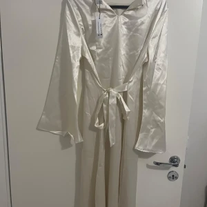 Champagnefärgad långklänning - Elegant champagnefärgad långklänning i satin med långa ärmar och ett knytband i midjan. Perfekt för festliga tillfällen eller en lyxig kväll. Klänningen har en enkel och stilren design som ger en sofistikerad look.