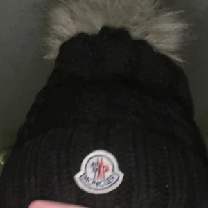 Svart stickad mössa med pälsboll - Säljer en snygg svart stickad mössa från Moncler med en fluffig pälsboll på toppen. Perfekt för kalla vinterdagar! Mössan har en klassisk design med Moncler-loggan framtill. Supermysig och stilren, passar till allt! 🖤