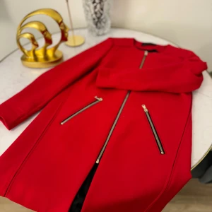 Röd kappa med dragkedjor - Snygg röd kappa i mycket bra skick med coola dragkedjor framtill. Perfekt för höst och vår, den har en stilren design utan krage och långa ärmar. Passar både till vardags och festliga tillfällen. En riktig färgklick i garderoben!