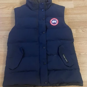 Blå canada goose väst - Hej Säljer en blå dunväst från Canada Goose i mycket bra skick. Västen har knappar och en klassisk logga på bröstet. Perfekt för kyliga höst- och vinterdagar. Den är både stilren och funktionell, med en normal passform och priset kan diskuteras.