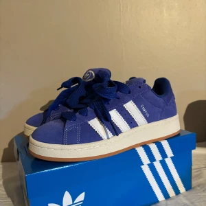 Blå Adidas Campus sneakers - Säljer ett par snygga blå Adidas Campus sneakers i mocka. De har de klassiska vita ränderna på sidan och kommer med både blå och vita skosnören. Skorna är i mycket bra skick (använda en gång)  och kommer i originalkartong, med vita och blåa snören. Köpta för 1 349 på Adidas egna hemsida