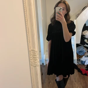 Svart sammet klänning - Säljer en elegant svart sammet klänning i mycket bra skick. Den har korta ärmar och en smockad detalj vid halsen som ger en fin textur. Perfekt för höst och vinter tillfällen. Klänningen är knälång och passar både till vardag och fest.