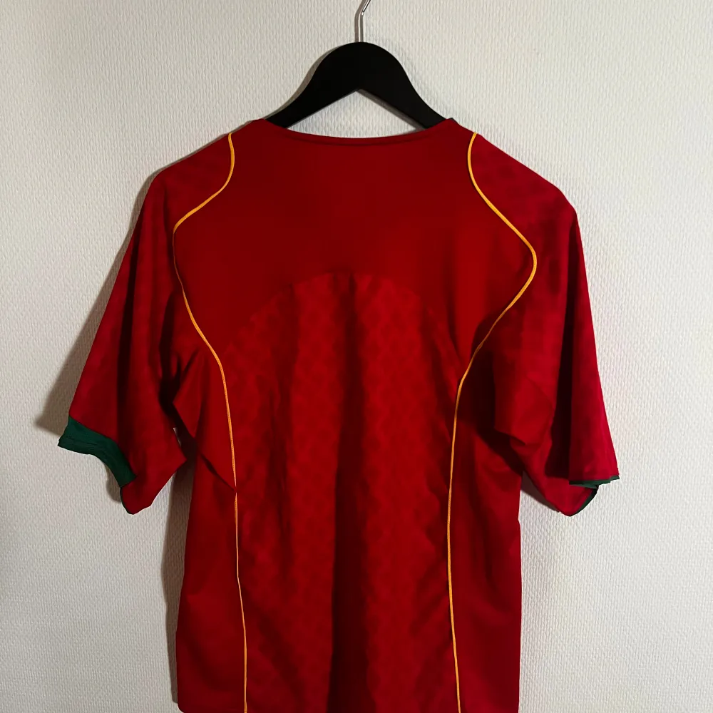 Säljer en röd Portugal fotbollströja från 2004 Nike i bra skick. Tröjan har korta ärmar och gröna detaljer vid kragen och ärmsluten. Det finns gula linjer längs sidorna och Portugals emblem på bröstet. Perfekt för fotbollsfans! ⚽. Sportswear.