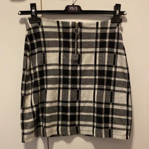 Rutig kjol från Lindex - Snygg rutig kjol från Lindex i svart och grått. Kjolen har en dragkedja framtill med en cool ringdetalj. Perfekt för både vardag och festliga tillfällen. Passar barn i åldern 10-12 år.