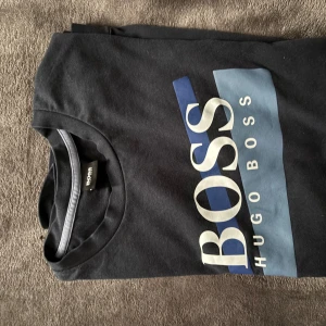 Svart t-shirt från Hugo Boss - Säljer en snygg svart t-shirt från Hugo Boss med stort tryck i vitt och blått på framsidan. Perfekt för en casual look eller att ha under en jacka. T-shirten är i bra skick och passar till både vardag och fest.