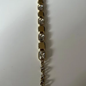 Guldarmband med klara stenar - Elegant guldarmband med rektangulära klara stenar infattade i varje länk. Perfekt för att ge en touch av glamour till vilken outfit som helst. Armbandet har en klassisk och tidlös design som passar både till vardags och festliga tillfällen.