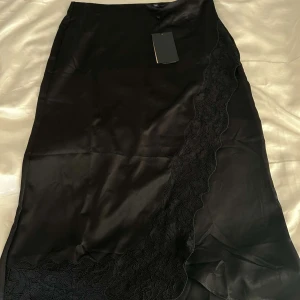 Svart sidenkjol med spetsdetaljer - Elegant svart långkjol i siden med vacker spetsdetalj längs ena sidan. Perfekt för festliga tillfällen eller en stilfull kvällslook. Kjolen har en mjuk och glansig yta som ger en lyxig känsla.