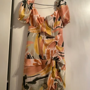 Färgglad klänning med volanger - Säljer en superfin klänning med färgglatt mönster i rosa, gul och svart. Den har korta puffärmar och volanger som ger en lekfull look. Perfekt för sommarens fester eller en dag på stan. Klänningen är i bra skick och passar perfekt för en solig dag!