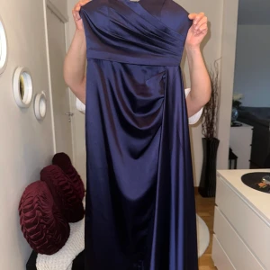 Mörkblå långklänning i satin - Elegant mörkblå långklänning i satin med omlott-detaljer upptill. Perfekt för festliga tillfällen som bal. Klänningen har en vacker glans och en smickrande passform.