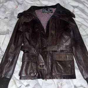 Brun skinnjacka med bälte - Säljer en snygg brun skinnjacka med bälte i midjan och dragkedja framtill. Jackan har långa ärmar och en cool rutig insida. Perfekt för höst och vår när du vill se stilren ut. Passar både till vardags och fest! Pris kan diskuteras!