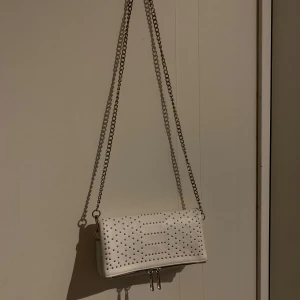 Vit axelväska med nitar - Snygg vit axelväska med coola nitar och kedja som axelrem. Perfekt för en utekväll eller fest! Väskan har en stilren design med tryckknappar för enkel öppning och stängning.