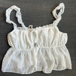 Vit ärmlös topp med volangdetaljer - Söt vit topp med volangdetaljer och små prickar. Den är ärmlös med smala axelband och har en luftig passform. Perfekt för sommardagar eller en avslappnad fest. Toppen har en knytning framtill för extra stil.