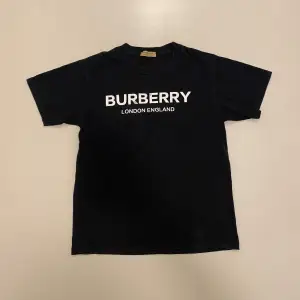 Säljer en snygg svart t-shirt från Burberry med vit text 'BURBERRY LONDON ENGLAND' på framsidan. Den är i bra skick och perfekt för en casual look. Passar både till vardags och fest. Storleken är inte angiven men ser ut att vara normal passform.