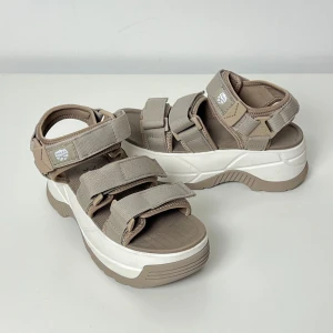 Shaka Sandaler mörk Beige Storlek 37 - Sandaler Neo Bungy Chunky SK-114 Beige.  Snygga och bekväma Shaka sandaler i beige färg. Perfekta för sommaren!  Denna sko kan passa alla typer av vriststorlek, och du kan justera passformen med hjälp av kardborrebanden.  Bekväm läst. Aldrig använd då storleken inte passade. Längd 24 cm storlek 37 Skons färg är mörkbeige, se bilderna.  Orginal pris 2400 kr
