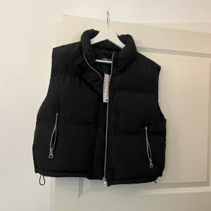 Svart dunväst med dragkedja - Säljer en snygg svart dunväst i mycket bra skick. Västen har en hög krage och praktiska dragkedjor på fickorna. Perfekt för höst och vinter när du vill hålla dig varm och stilren. Passar både till vardags och mer avslappnade tillfällen.