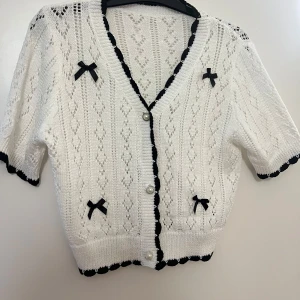 Vit stickad kofta med svarta detaljer - Superfin vit stickad kofta med svarta kanter och söta rosetter. Den har en v-ringad design och knäpps med pärlknappar. Perfekt för vår och sommar, och ger en gullig touch till vilken outfit som helst. Ärmarna är trekvarts och koftan har ett fint mönster.