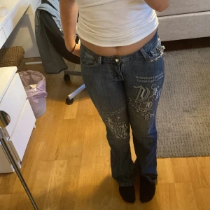 Blå jeans med tryck - Säljer ett par blå jeans med coolt tryck på framsidan. De är lågmidjade och har en bootcut still. Jeansen har slitna detaljer på bakfickorna och är perfekta för en avslappnad stil.