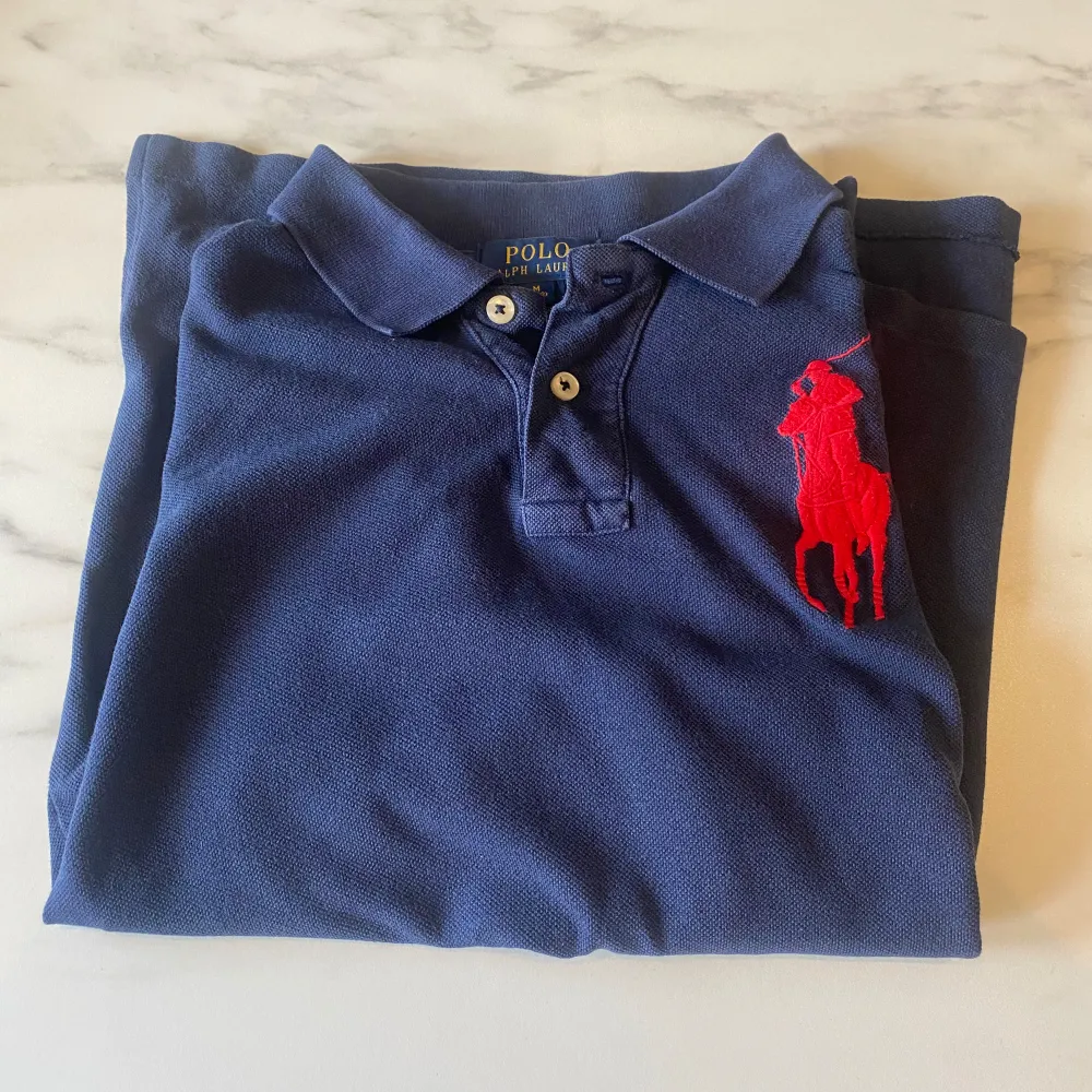 Säljer en snygg mörkblå pikétröja från Ralph Lauren med det klassiska röda broderade logotypen på bröstet. Tröjan har korta ärmar och en stilren krage med knappar. Perfekt för en casual look eller en dag på stan! Är liten i storleken så passar även xs och s.. T-shirts.