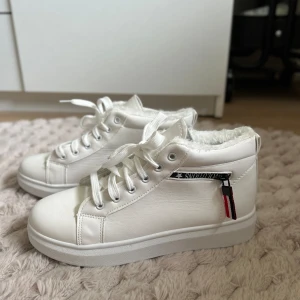 Vita sneakers med foder och dragkedja - Säljer ett par vita sneakers med mysigt foder inuti, perfekt för kyligare dagar. Skorna har snörning framtill och en cool dragkedja på sidan med en liten röd och svart detalj. De är i nyskick och redo att användas direkt!