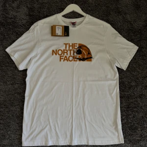 Vit t-shirt från The North Face - Säljer en snygg vit t-shirt från The North Face med deras ikoniska logga i orange på bröstet. T-shirten är i nyskick och har korta ärmar. Perfekt för en avslappnad stil eller för att bära under en hoodie.