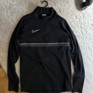 Säljer en svart Dri-FIT tröja från Nike i storlek S. Den har en stilren design med en vit Nike-logga på bröstet och en diskret dragkedja vid halsen. Perfekt för träning eller vardagsbruk. Materialet är lätt och andas bra, vilket gör den idealisk för aktiva dagar.