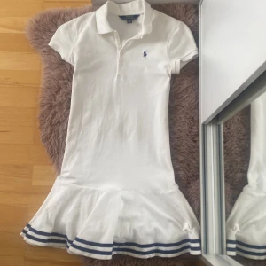 Vit klänning från Ralph Lauren - Säljer en superfin vit klänning från Ralph Lauren. Den har  pikékrage och korta ärmar. Kjolen har en söt volangkant med blå ränder längst ner. Perfekt för sommaren. helt oanvänd!💕storlek 12-14år