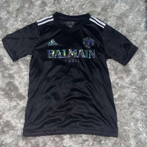 Svart fotbollströja från Adidas x Balmain - Säljer en snygg svart fotbollströja från samarbetet mellan Adidas och Balmain. Tröjan har korta ärmar och de klassiska tre ränderna på axlarna. Den är i nyskick och har en cool, skimrande Balmain-logga på bröstet. Perfekt för både träning och vardag!