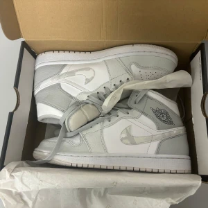 Air Jordan MID camo grey - Ett par vita och gråa air Jordan 1 MID i storlek eu 40,5 för män med camoflage detaljer på swooshen inhandlade 2020. Är använda, men bra skick (se bilder) kan visa kvitto om så önskas!