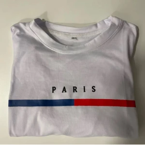 Vit t-shirt med Paris-tryck - Säljer en snygg vit t-shirt med texten 'PARIS' tryckt på framsidan. Den har en enkel design med en blå och röd linje under texten. Perfekt för en casual look!