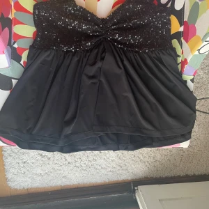 Svart paljettprydd topp - Säljer en snygg svart topp med paljetter upptill. Toppen har en glittrig överdel med ett knytliknande mönster i mitten och ärmlös design. Perfekt för festliga tillfällen!