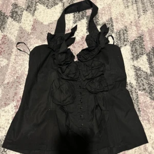 Svart korsett med volangdetaljer - Säljer en snygg svart korsett med volangdetaljer framtill. Den har en halterneck-design och knappar längs framsidan. Perfekt för en festlig kväll eller en speciell tillställning. Passar bra till både kjol och byxor!