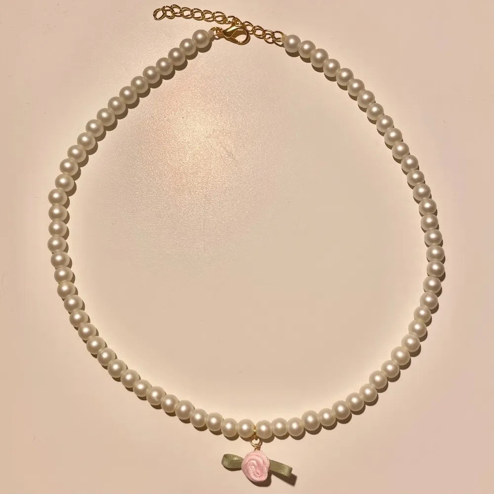 Säljer ett superfint handgjort pärlhalsband med vita pärlor och en söt rosa rosett som hänge. Halsbandet har en justerbar kedja i guld för perfekt passform. Perfekt för att ge en elegant touch till vilken outfit som helst!. Accessoarer.