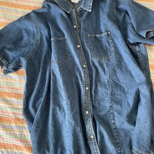 Blå jeansskjorta från Duo Maternity - Säljer en snygg blå jeansskjorta från Duo Maternity. Skjortan är kortärmad och har knappar hela vägen fram. Den är tillverkad i 100% bomull vilket gör den både bekväm och hållbar. Perfekt för en avslappnad stil!