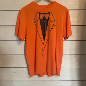 Orange t-shirt från Off-White x Nike - Säljer en cool orange t-shirt från Off-White x Nike. Den har ett tryck av en smoking på framsidan och en stor örn med 'LOGO' och Nike-swoosh på baksidan. Perfekt för dig som vill sticka ut! T-shirten är i bomull och har korta ärmar. Aldrig använd 