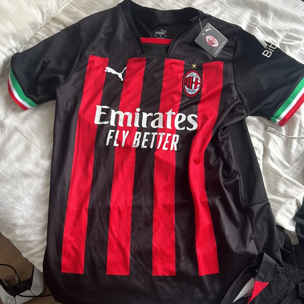 Säljer en snygg AC Milan fotbollströja från Puma. Den är svart med röda ränder och har korta ärmar med italienska flaggans färger på muddarna. Tröjan har lagets emblem och sponsorlogga på framsidan. Perfekt för alla Milan-fans! helt ny med tags och alldrig använd. T-shirts.